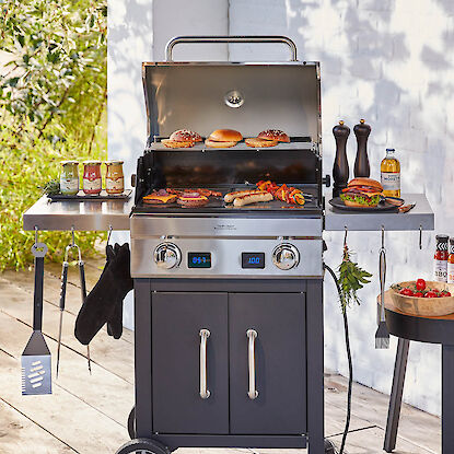 Gasgrill mit Burgern, Würstchen und Beilagen, umgeben von Grillzubehör, Saucen und Gewürzen
