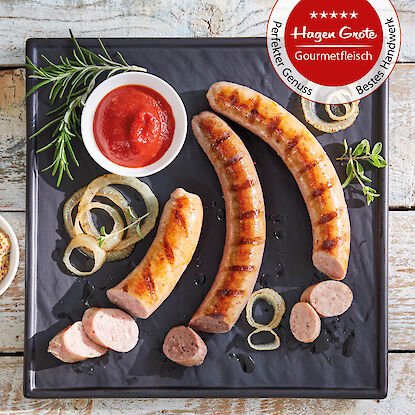 Gegrillte Bratwürste mit Zwiebeln, Kräutern und Ketchup auf schwarzem Teller, Siegel „Gourmetfleisch“.