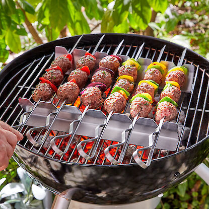 Grillspieße mit Hackfleisch und Gemüse über heißer Glut auf rundem Holzkohlegrill im Garten.