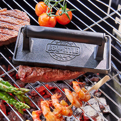 Steak mit Grillpresse, Garnelen-Spieße, Spargel und Tomaten auf Grillrost über heißer Glut.