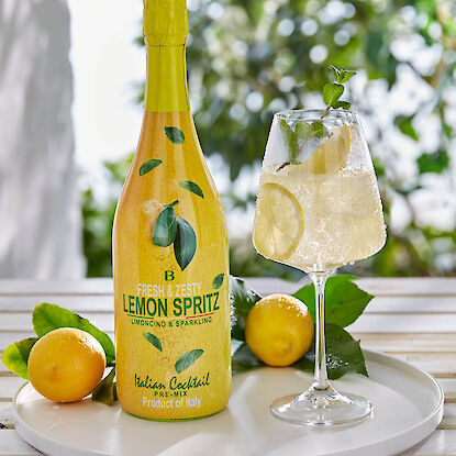 Flasche Lemon Spritz mit gefülltem Cocktailglas, Zitronenscheiben und Minze auf weißem Tablett.