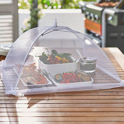 Speisenschutzhaube über Schalen mit Grillgemüse und Garnelen auf Holztisch neben Gasgrill im Garten.