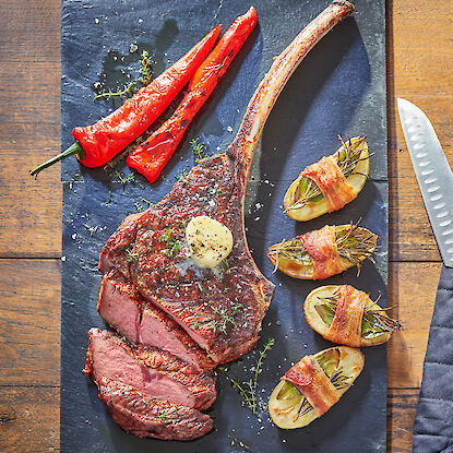 Tomahawk-Steak mit Kräuterbutter, gegrillten Paprika und Speck-Kartoffeln auf dunkler Schieferplatte.