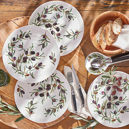 Teller mit Olivenzweig-Motiv, Besteck, Brotschale und Deko auf rustikalem Holztisch mediterran gedeckt.
