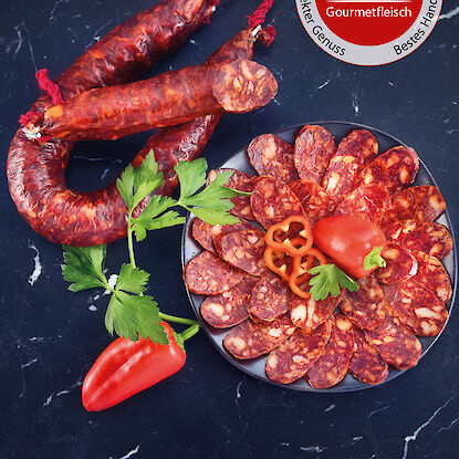 Chorizo-Rohwurst im Ganzen und in Scheiben angerichtet, mit Paprika, Petersilie und Gourmetfleisch-Siegel.