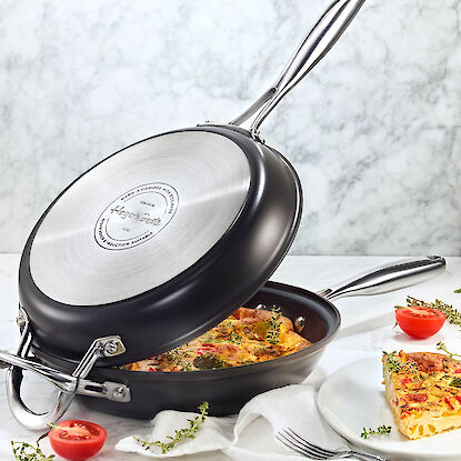Doppelfunktionale Pfanne mit Gemüse-Frittata, angerichtet mit Kräutern und Tomaten auf Marmorplatte.