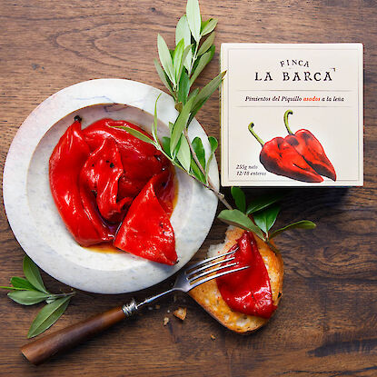 Geröstete Piquillo-Paprika auf Brot und in Schale, daneben Originalverpackung und Olivenzweige auf Holztisch.