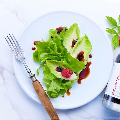 Salat mit Himbeeren und Vinaigrette aus Vincotto Lampone, daneben Flasche und frische Himbeerblätter.