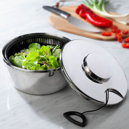 Edelstahl-Salatschleuder mit frischem Blattsalat, daneben Schneidebrett mit Messer, Paprika und Frühlingszwiebeln.
