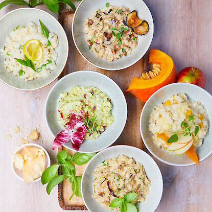 Sechs Schalen mit verschiedenen Risotto-Variationen, dekoriert mit frischen Zutaten auf hellem Holztisch.