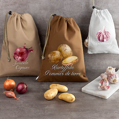 Stoffbeutel für Zwiebeln, Kartoffeln und Knoblauch mit Aufdruck und Gemüse, dekorativ arrangiert.
