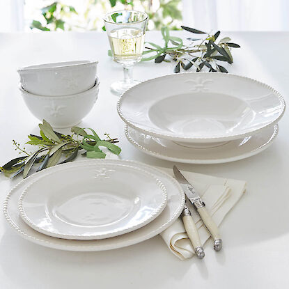 Weißes Geschirr mit Reliefdekor, Glas mit Weißwein und Besteck auf elegant gedecktem Tisch mit Olivenzweigen.