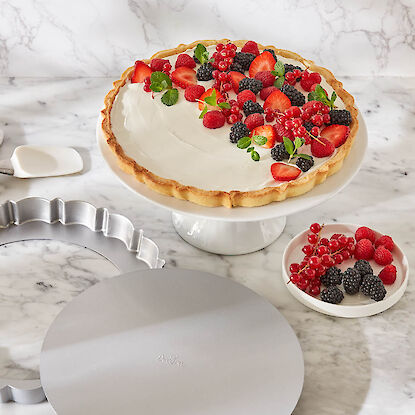 Beerentarte mit Sahne auf Tortenplatte, daneben Backform, Beeren und Tortenheber auf Marmorfläche.