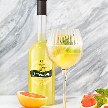 Limoncello-Flasche mit gefülltem Cocktailglas, Grapefruitstücken und Minze auf hellem Untergrund.