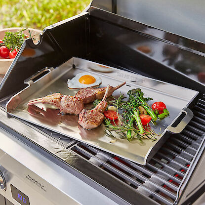 Gasgrill mit Grillplatte, darauf Lammkoteletts, Gemüse, Kräuter und Spiegelei beim Braten.