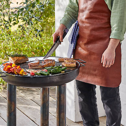 Mann am Grill mit Leder-Schürze grillt Fleisch und Gemüse auf runder Feuerstelle auf der Terrasse.