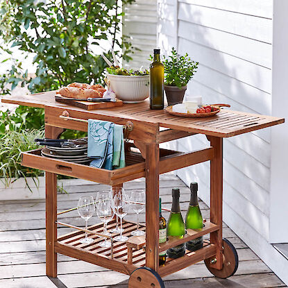 Servierwagen aus Holz mit Geschirr, Gläsern, Brot, Salat, Wein und Kräutern auf Terrasse im Freien.