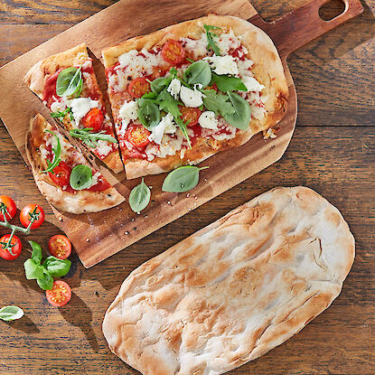 Fladenbrot mit Tomaten, Käse und frischen Kräutern auf Holzbrett, daneben ungebackenes Brot und Deko.