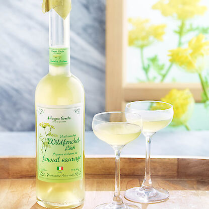 Flasche Wildfenchel-Likör mit zwei Gläsern auf Tablett vor blumigem, hellem Hintergrund.