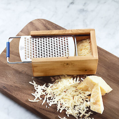 Käsereibe aus Holz mit geriebenem Käse und Parmesanstücken auf Holzbrett und Marmorfläche.