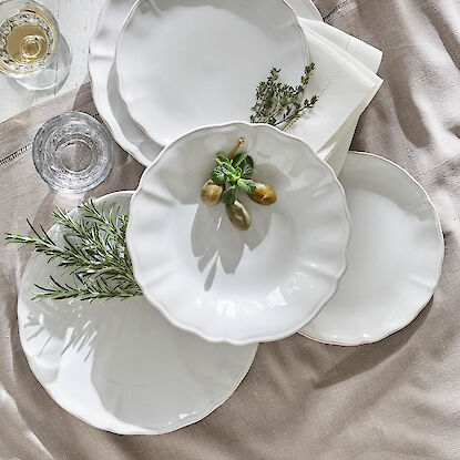 Elegante weiße Teller mit frischen Kräutern und Gläsern auf beigefarbener Tischdecke.