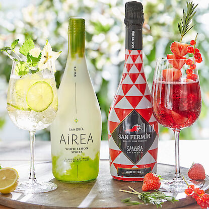 Zwei Sangría-Flaschen mit passenden Sommerdrinks, garniert mit Früchten und Kräutern auf einem Holztablett.