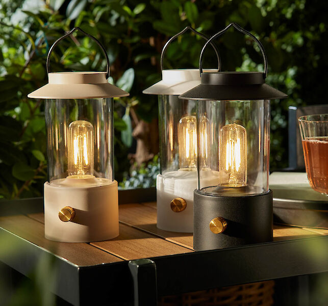 Drei stilvolle LED-Laternen mit Glaskörpern leuchten auf einem Gartentisch im Abendlicht.