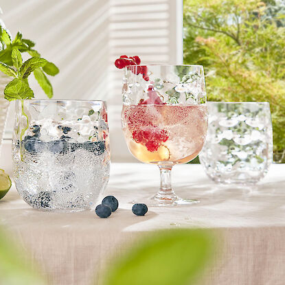 Geprägte Trinkgläser mit Sommergetränken, Beeren und Minze auf hell gedecktem Tisch.