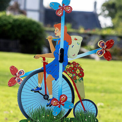 Bunte Garten-Windmühle mit Radfahrerin, Schmetterlingen und Blumen auf grüner Wiese.