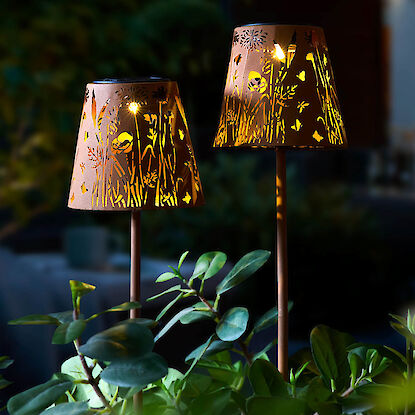 Zwei leuchtende Gartenlampen mit Naturmotiven im Dunkeln zwischen grünen Pflanzen.