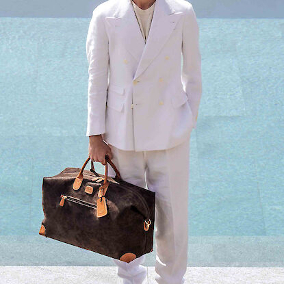 Mann in weißem Anzug mit brauner Reisetasche steht vor Pool in sonniger Umgebung.