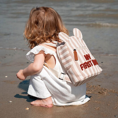 Kleines Kind mit gestreiftem Rucksack „My First Bag“ spielt barfuß am Strand.