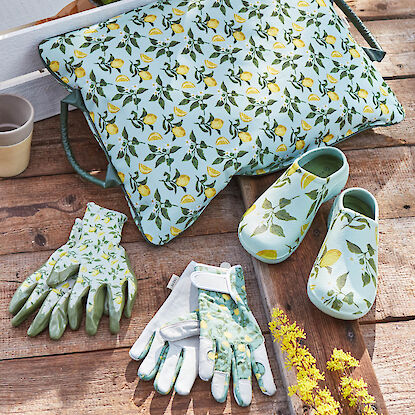 Gartenhandschuhe, Schuhe und Kniekissen mit Zitronenmotiv auf Holzboden im Sonnenlicht.