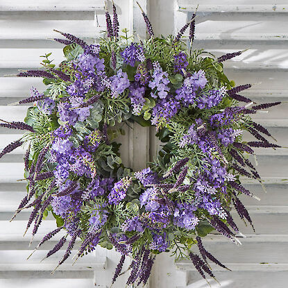 Kranz aus violetten Kunstblumen und Grün an weißer Holzjalousie aufgehängt.