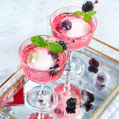 Zwei rosa Cocktails mit runden Eiswürfeln, Brombeeren und Basilikum auf Spiegeltablett.