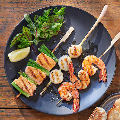 Grillspieße mit Garnelen, Jakobsmuscheln und Lachs auf dunklem Teller mit Salat und Zitrone.