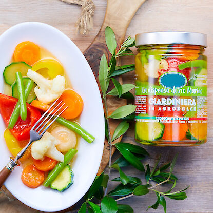 Glas Giardiniera-Gemüse mit bunter Gemüseplatte, Gabel und Olivenzweigen auf Holztisch.