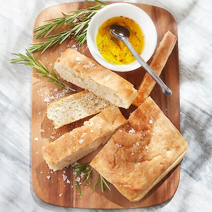 Focaccia mit Meersalz auf Holzbrett, dazu Olivenöl mit Kräutern und Rosmarinzweige.