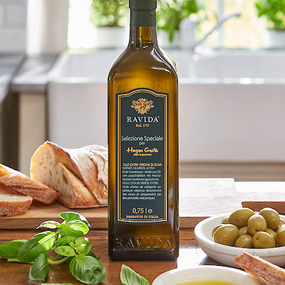 Flasche Olivenöl mit Brot, Oliven, Basilikum und Öl-Schälchen auf Holzplatte in Küche.