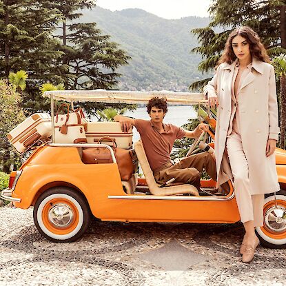 Paar mit Retro-Gepäck in orangefarbenem Cabrio vor Palmen und See in sommerlicher Kulisse.