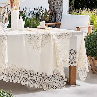Cremefarbene Spitzentischdecke auf Gartentisch mit Deko und Sitzgruppe im Grünen.