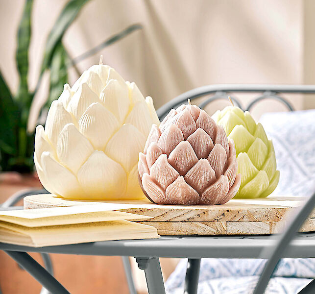 Drei Kerzen in Artischockenform in Creme, Rosa und Grün auf Holzbrett auf Gartentisch.