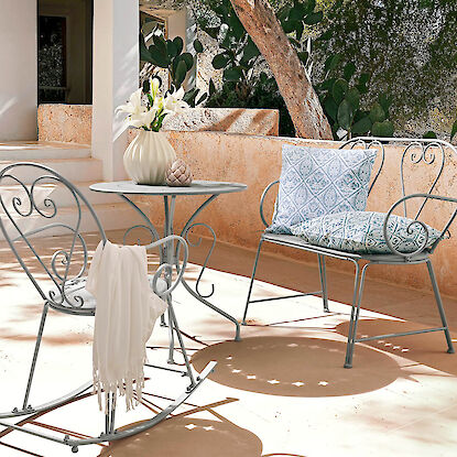 Metallmöbel mit gemusterten Kissen auf Terrasse, kleiner Tisch mit Vase und Schaukelstuhl.