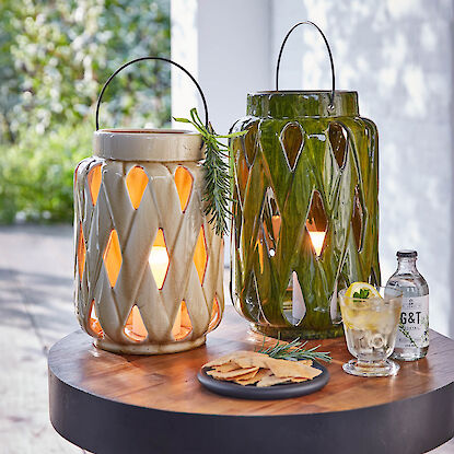 Zwei Keramiklaternen mit Licht auf Holztisch, dazu Drink, Cracker und Rosmarinzweig.
