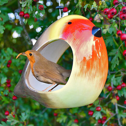 Rotkehlchen mit Wurm sitzt in bunter Vogel-Futterstelle aus Keramik, umgeben von Beerenzweigen.