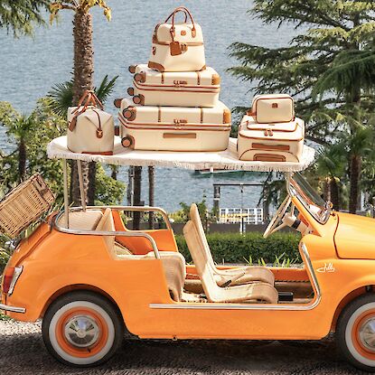 Paar mit Retro-Gepäck in orangefarbenem Cabrio vor Palmen und See in sommerlicher Kulisse.