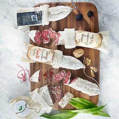 Sélection de saucissons français emballés dans du papier, présentés sur une planche en bois avec tranches coupées, noix, baies roses et herbes.