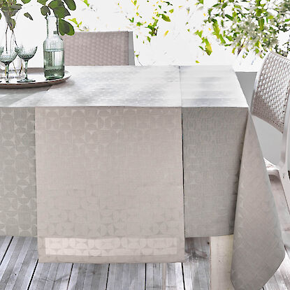 Nappe grise à motif géométrique discret avec chemin de table assorti, verres et carafe sur un plateau, table dressée en extérieur.