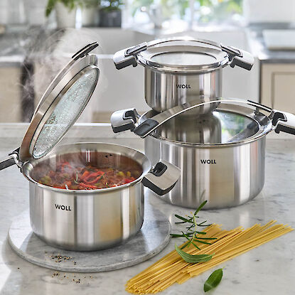 Trois casseroles en inox de la marque Woll, l’une avec un ragoût fumant, des spaghettis crus et des herbes fraîches posés sur un plan de travail en marbre.