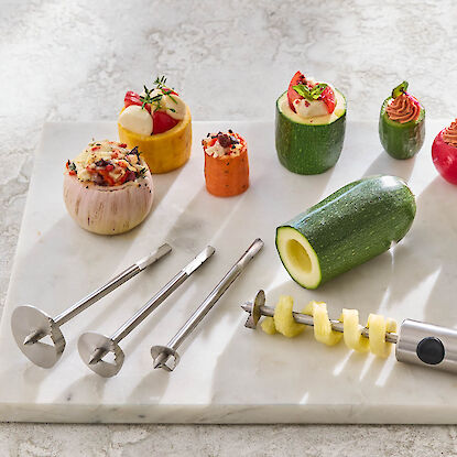 Coupe-légumes avec différents embouts, courgettes, poivrons, oignon et carotte évidés et farcis, présentés sur une plaque en marbre.